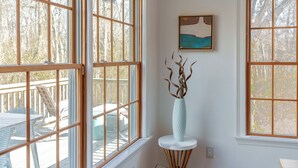 Ein Zimmer mit großen Fenstern, einer weißen Vase mit Zweigen und einem Gemälde an der Wand.