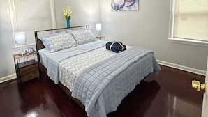 A bedroom with a bed, bedside tables, a nightstand, a window with blinds, a wall-mounted artwork, and a ceiling light.