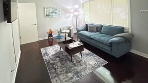 A living room with a dark wood floor, a blue sofa, a coffee table, a rug, a TV, and a window with blinds.
