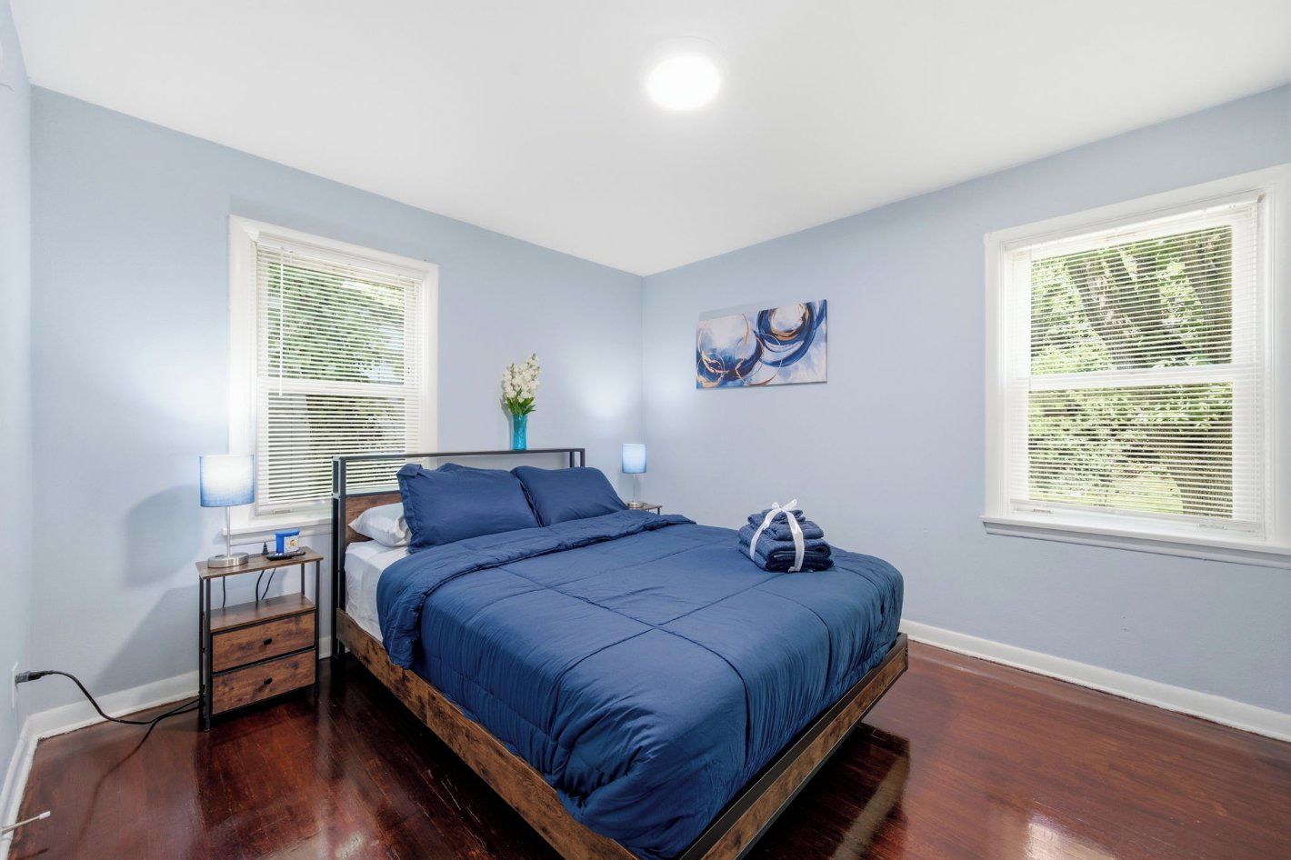 Un dormitorio con cama, mesitas de noche, lámpara, un jarrón con flores y un cuadro en la pared.