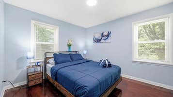 Un dormitorio con cama, mesitas de noche, lámpara, un jarrón con flores y un cuadro en la pared.