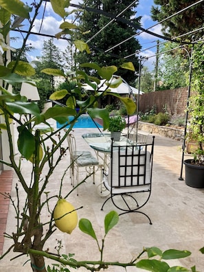 A patio area with a glass table and chairs, a small lemon tree, and a pool in the background. - Beautiful, spacious villa—perfect for a summer vacation for 5–6 people - (Le Rouret)