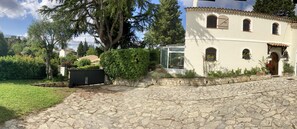 A stone-paved courtyard with a glass greenhouse, a white building with arched windows, and surrounding greenery. - Beautiful, spacious villa—perfect for a summer vacation for 5–6 people - (Le Rouret)