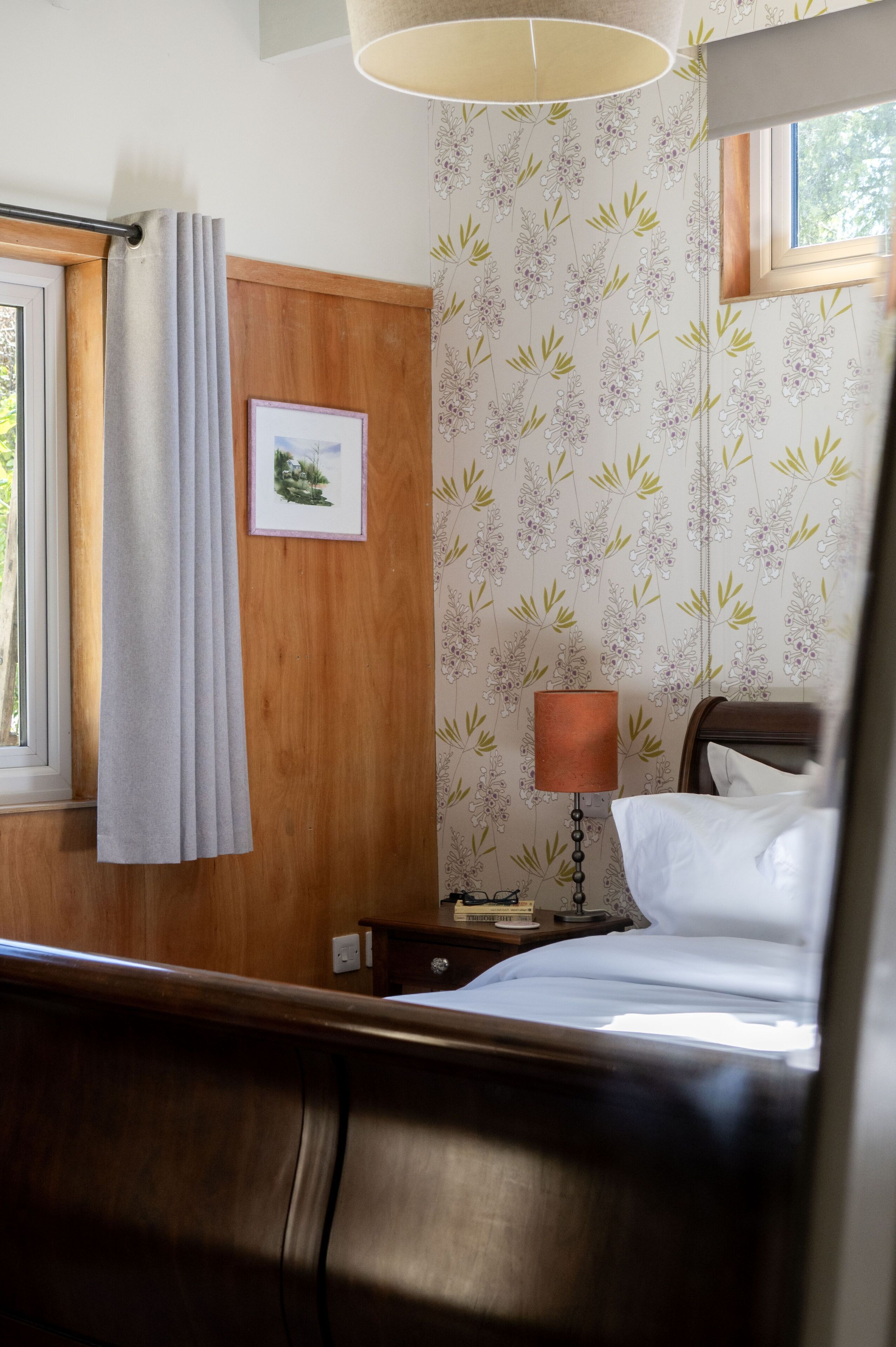 A hotel room with a wooden headboard, a bed with white linens, a nightstand with a lamp, a framed picture on the wall, and a window with a curtain.