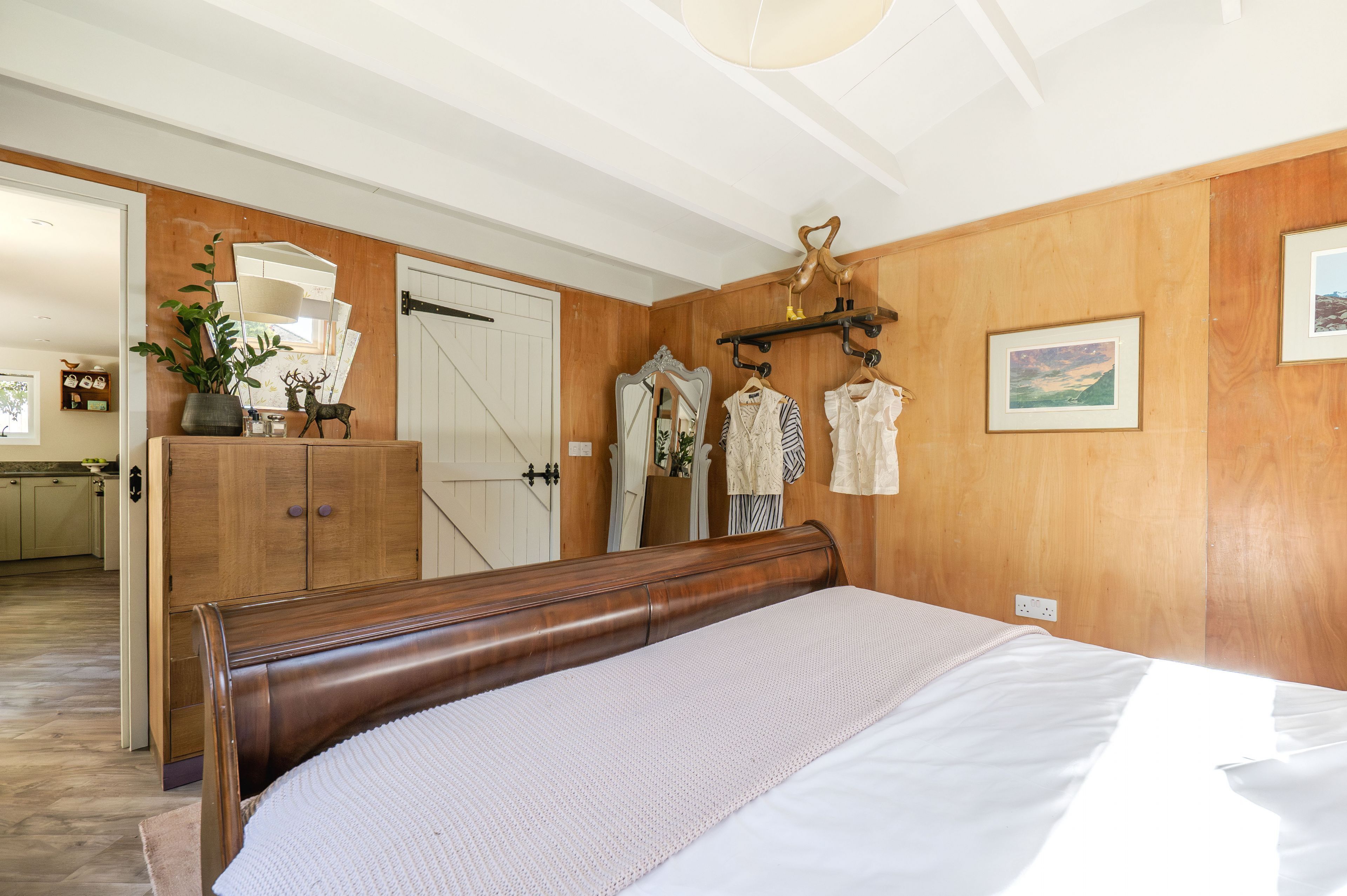 Une chambre à coucher avec une tête de lit en bois, un lit avec une literie blanche, une commode, un miroir et un porte-manteau avec des vêtements.
