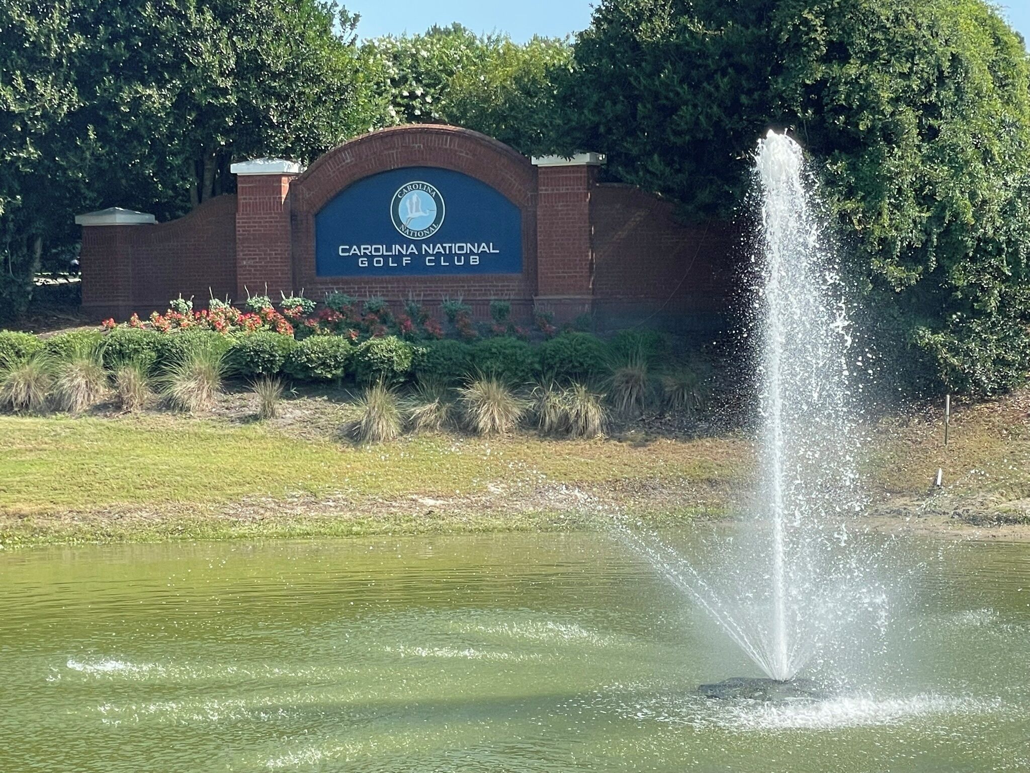 A sign for Carolina National Golf Club with a water fountain in the foreground.