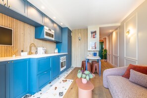 A modern kitchen with blue cabinets, a white countertop, and a wooden floor. There is a dining area with a table and chairs, and a living room with a sofa. - Waimea - À 200 m de la plage (Capbreton)