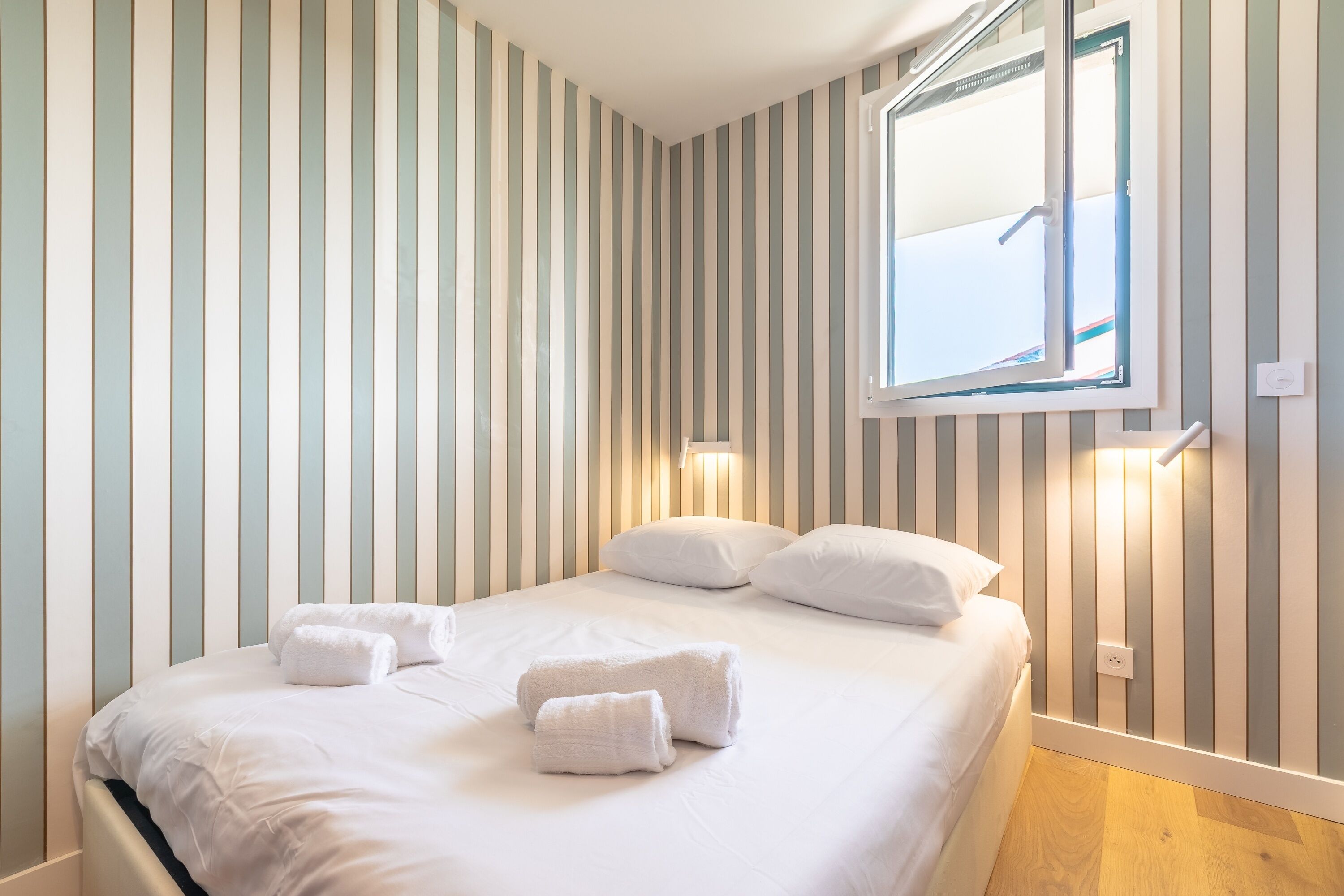 A neatly made bed with white linens and towels, a window with a view, and striped wall decor.