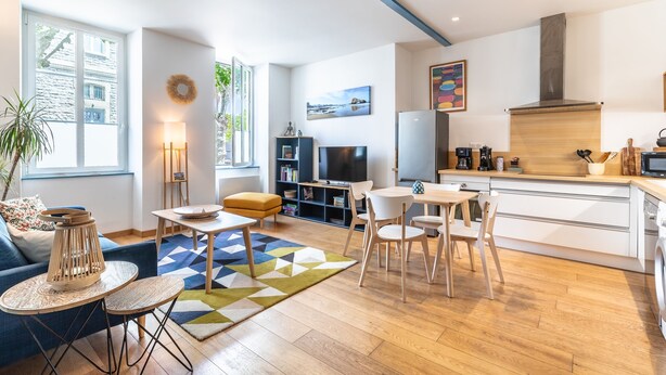 A modern living room with a sofa, coffee table, and TV, a kitchen with a stove and microwave, and a dining area with chairs and a table. - Daisy – Élégant cocon au cœur de Biarritz (Biarritz)