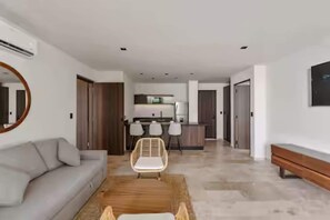 A modern living room with a sofa, a coffee table, and a dining area with chairs and a kitchen in the background. - Chic Jungle 2BR - Balcony, Priv Pool, Steam & Gym (Tulum)