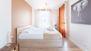 A bedroom with a bed, a nightstand, a wall-mounted light, a window with curtains, and a wall art of an elephant. - Carnet de route - Appt climatisé avec balcon (Lyon)