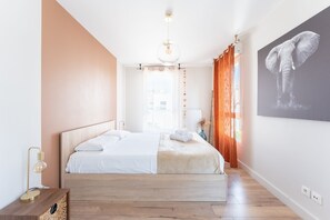 Ein Schlafzimmer mit einem Bett, einem Nachttisch, einer Wandlampe, einem Fenster mit Vorhängen und einem Wandbild eines Elefanten.