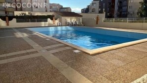 Ein rechteckiger Pool mit einer gefliesten Liegefläche, umgeben von einer niedrigen Mauer und einem Gebäude im Hintergrund.