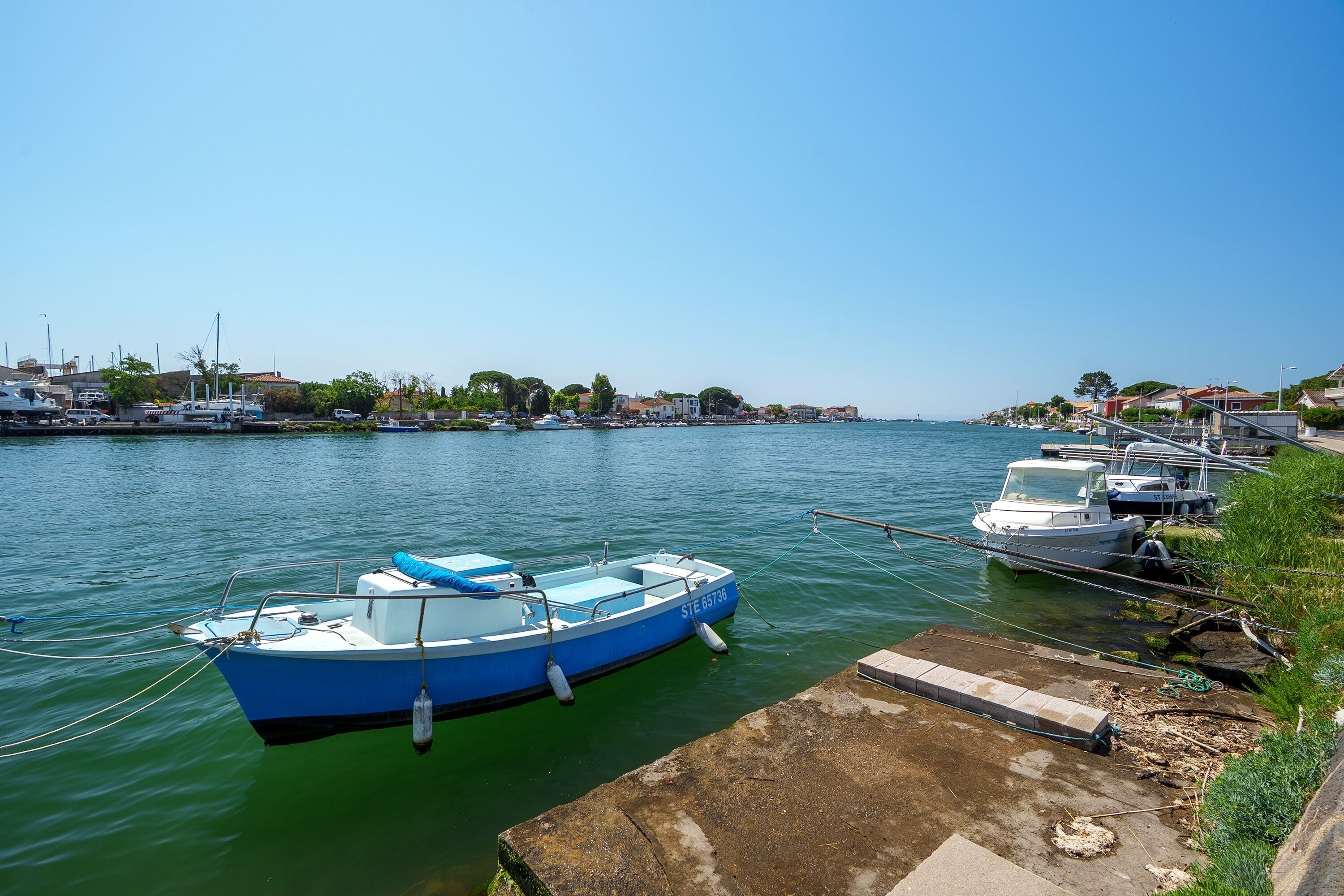 Uma marina com barcos atracados, céu claro e edifícios ao fundo.