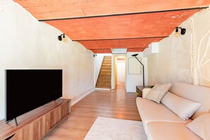 A living room with a flat-screen TV on a wooden stand, a white sofa with cushions, a wooden staircase, and a wall-mounted lamp. - Le Resticas - Triplex climatisé (Rustiques)