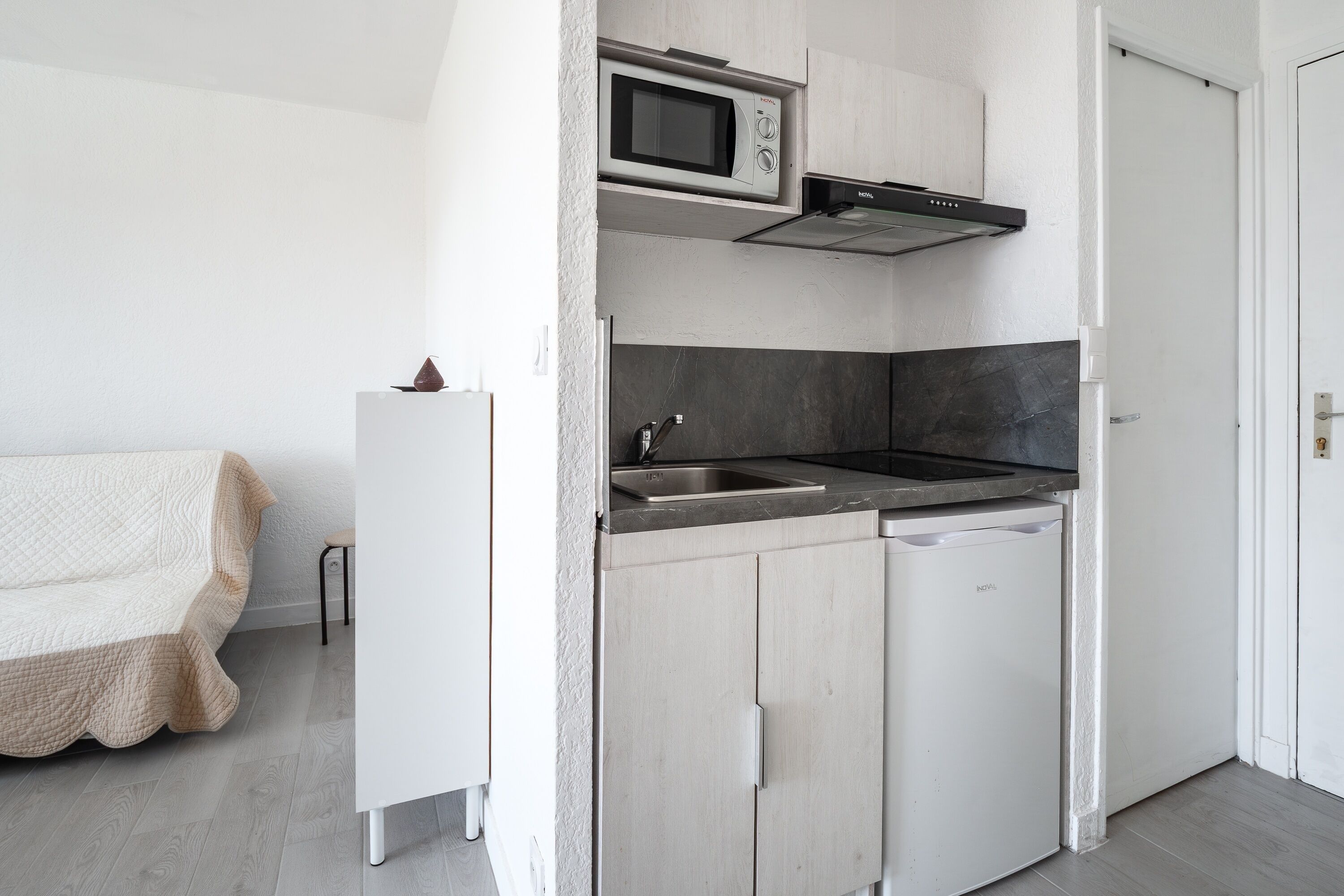 A compact kitchen with a microwave, sink, and dishwasher, a white sofa, and a door to the right.
