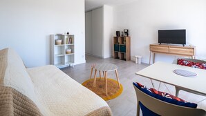 A modern living room with a sofa, a dining table, a TV, and a small stool. - Le Refuge de Canet - Studio climatisé (Canet-en-Roussillon)