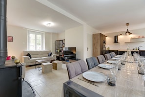 Ein modernes Wohnzimmer mit einer Couch, einem Couchtisch und einem Fernseher. Man sieht einen Essbereich mit einem langen Tisch, gedeckt für sechs Personen. Die Küche verfügt über Holzschränke und einen eingebauten Backofen.