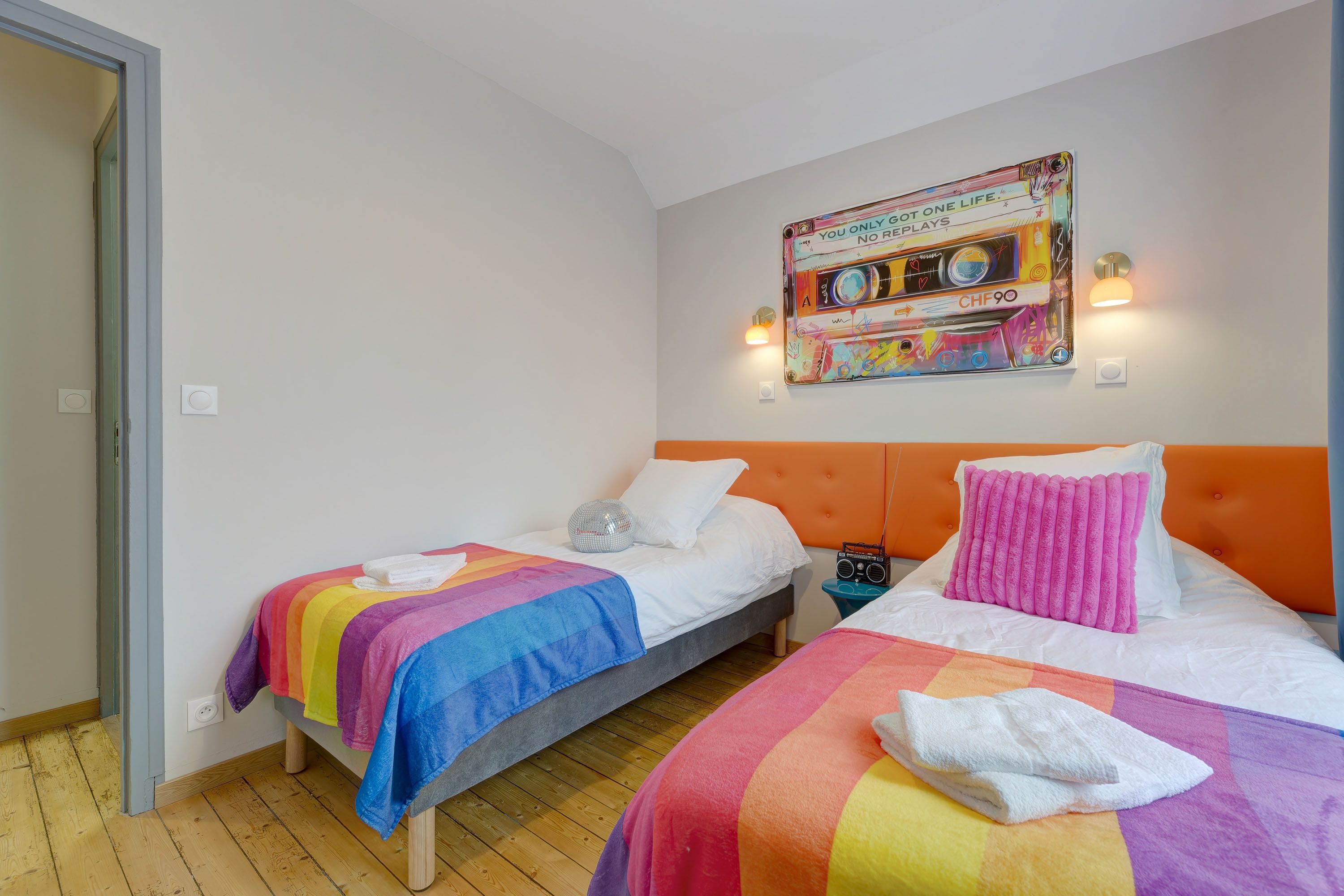 A hotel room with two single beds, a colorful wall art piece, a radio on the nightstand, and a wooden floor.