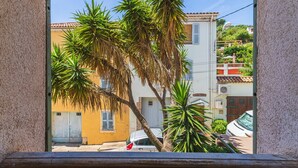 Une vue à travers la fenêtre d’un bâtiment, avec des palmiers, une voiture garée et un immeuble jaune avec des volets.