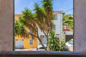 Blick durch ein Fenster auf ein Gebäude mit Palmen, ein geparktes Auto und ein gelbes Gebäude mit Fensterläden.