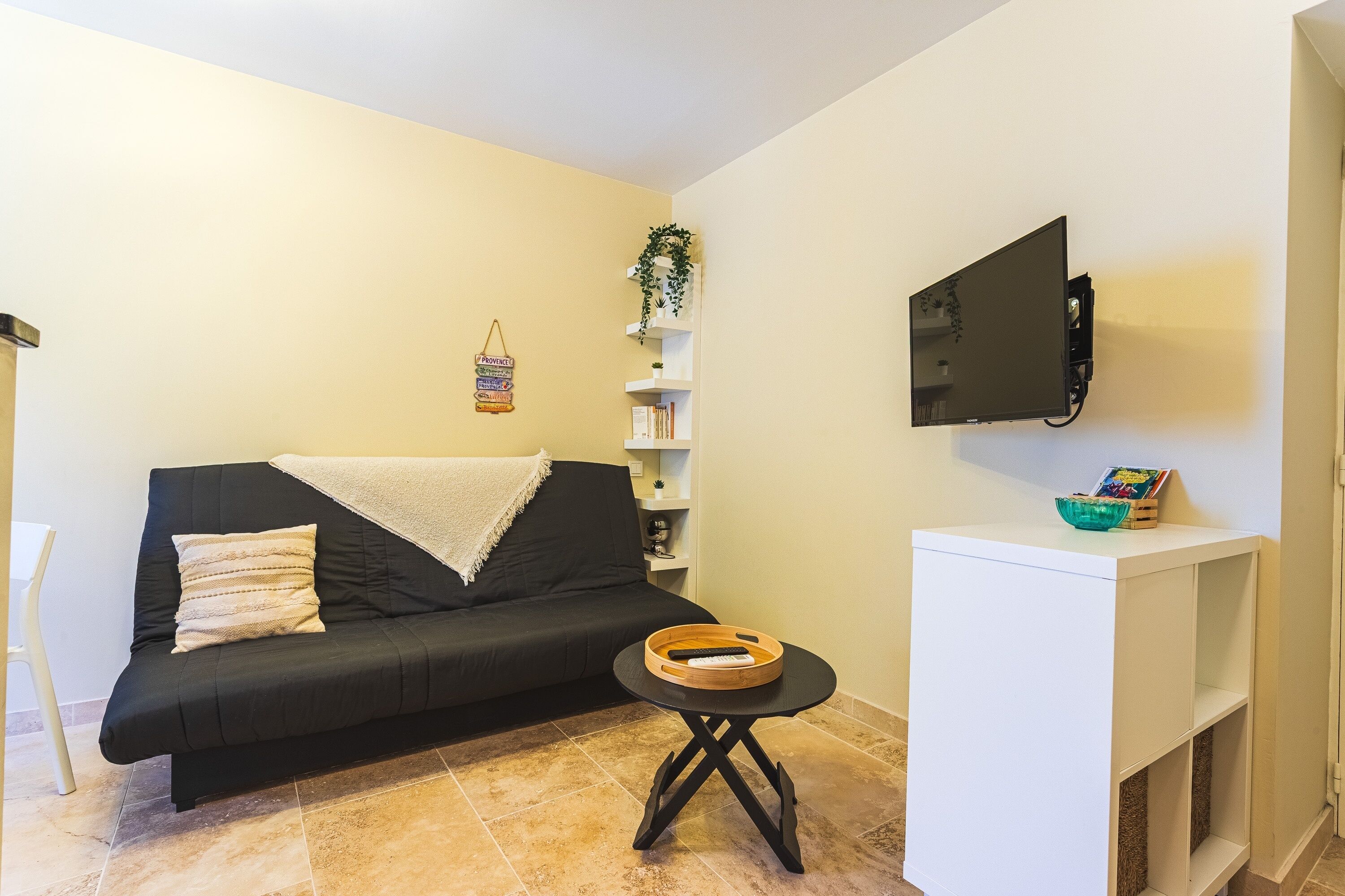 A living room with a black sofa, a small wooden side table, a white console table, a mounted TV, and a wall-mounted shelf with decorative items.
