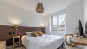 A bedroom with a large bed, bedside lamps, a wooden nightstand, a desk with a computer, and a window with a view. - Le Cocon des sables – climatisé à 100m de la plage (Asnelles)