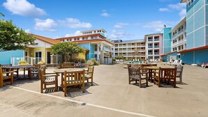 A hotel courtyard with outdoor seating, a pool, and multiple buildings with balconies. - Bayside Condo Steps to Beach with Resort Pools (Virginia Beach)