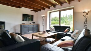 Ein Wohnzimmer mit Holzbalkendecke, einem schwarzen Ledersofa, einem Couchtisch und Blick durch ein großes Fenster.
