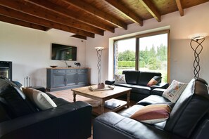 Ein Wohnzimmer mit Holzbalkendecke, einem schwarzen Ledersofa, einem Couchtisch und Blick durch ein großes Fenster.