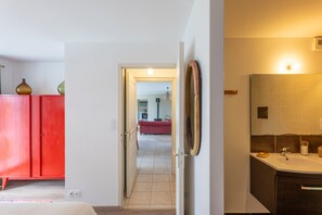 A hotel room with a red wardrobe, a bathroom with a mirror and sink, and a view into the living area. - La Ville Barbier - Belle maison avec piscine (Médréac)