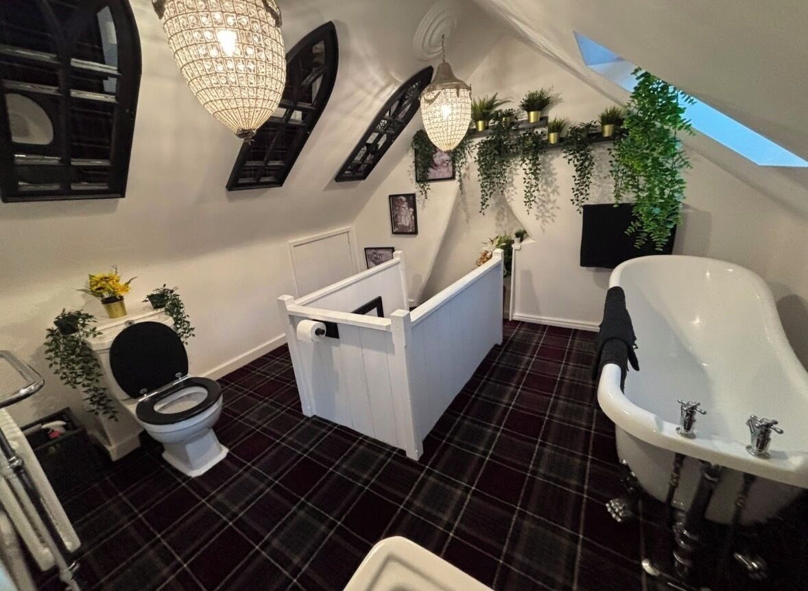 A bathroom with a white bathtub, a toilet, and a shower area. There are plants and a chandelier.
