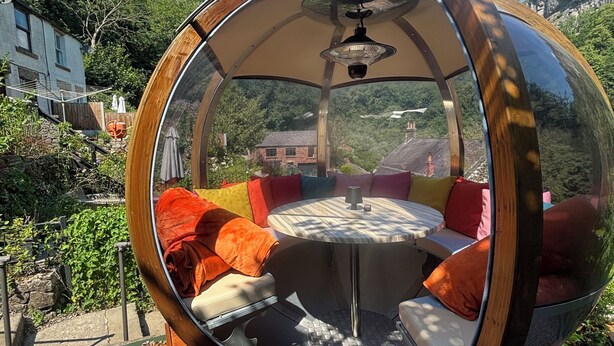 A spherical room with a round table and chairs, surrounded by cushions, set on a wooden deck with a view of a house and mountains. - Cliff View Cottage Holiday Let (Matlock Bath)