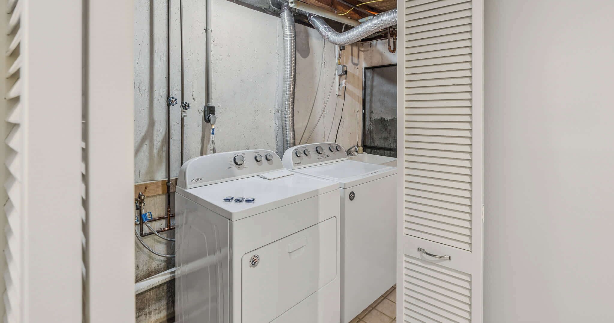A laundry room with two Whirlpool washers and dryers, exposed ductwork, and a wooden ceiling.