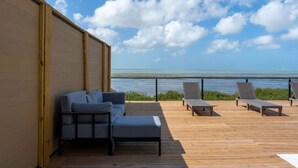 Eine Holzterrasse mit einer Couch und zwei Liegestühlen, mit Blick auf ein Gewässer.