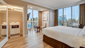 A hotel room with a large bed, a walk-in closet, a small table with chairs, and a view of the city through large windows.