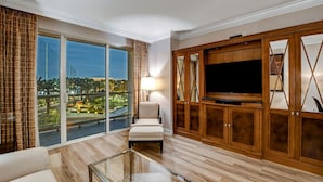 A spacious living room with a large TV, comfortable seating, a glass coffee table, and a view of a cityscape through large windows.