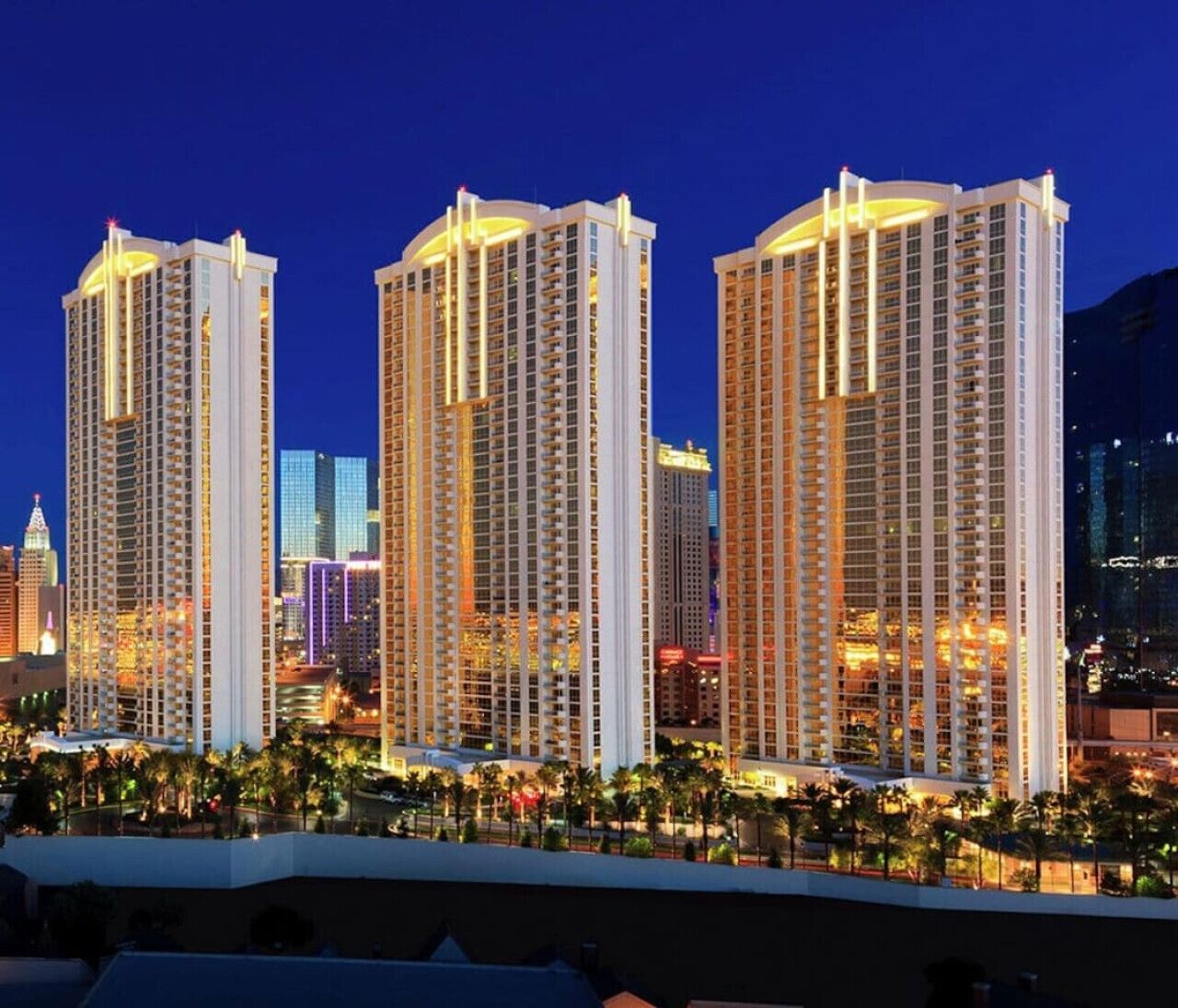 Three high-rise buildings with balconies, surrounded by palm trees and a well-lit urban skyline.