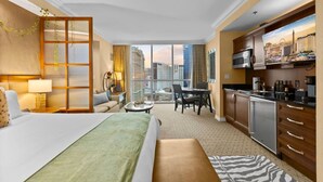 Ein Hotelzimmer mit einem Bett, einer Sitzecke mit Tisch und Stühlen, einer Küchenzeile und einem großen Fenster mit Blick auf die Stadt.