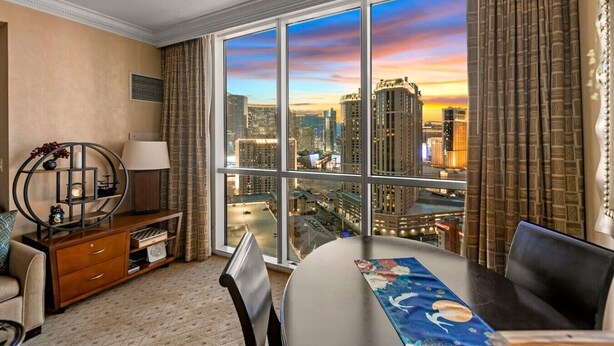 Ein Hotelzimmer mit großem Fenster und Blick auf die Stadt, einem runden Tisch, einem Sessel, einem Schreibtisch mit Lampe sowie einem dekorativen Regal.