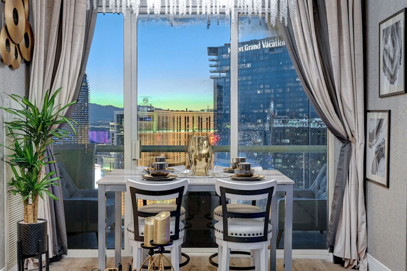 A dining area with a table set for two, a potted plant, and a view of a cityscape through large windows.