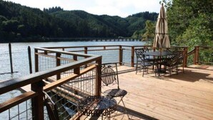 A wooden deck with a table and chairs overlooking a lake, surrounded by forested hills.