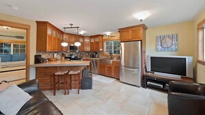 A modern kitchen with wooden cabinets, a stainless steel refrigerator, a dining area with a sofa, and a TV.