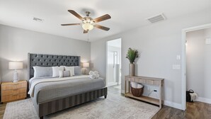 Une chambre à coucher avec une tête de lit capitonnée, un ventilateur de plafond, une table de chevet avec une lampe et une console avec un panier.
