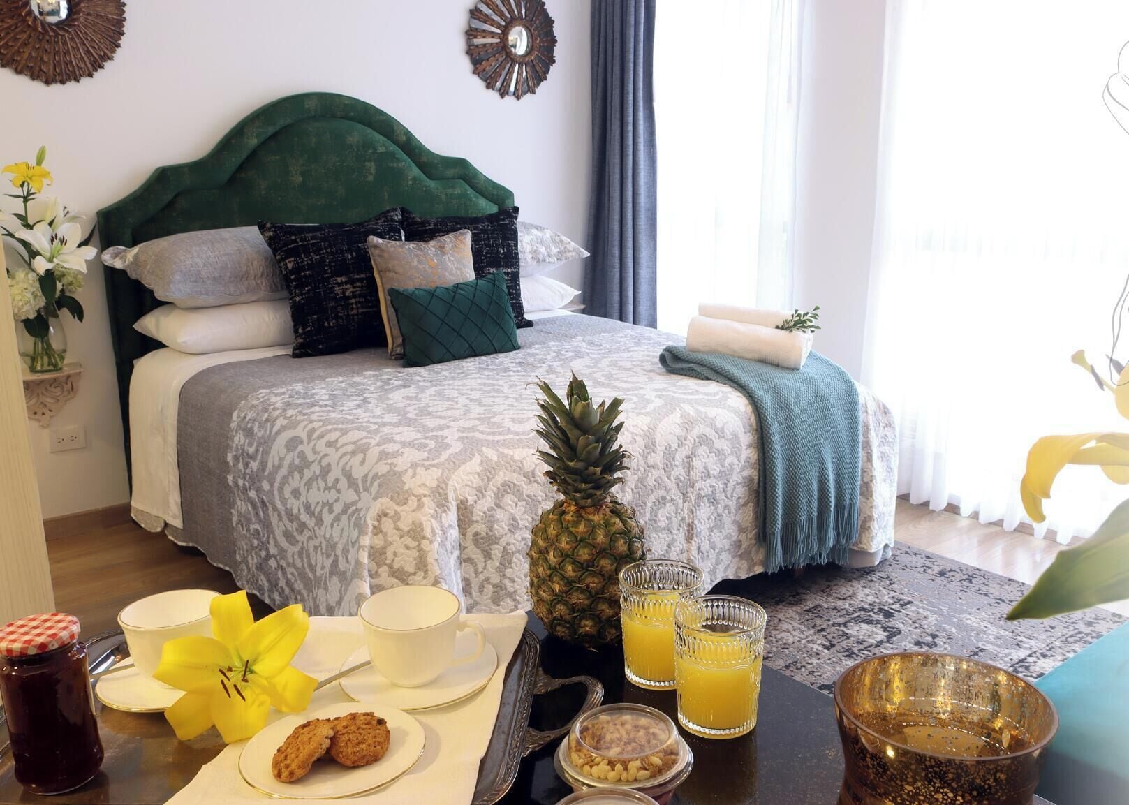 A neatly made bed with patterned bedding, a pineapple, breakfast items, and a view of a window with curtains.