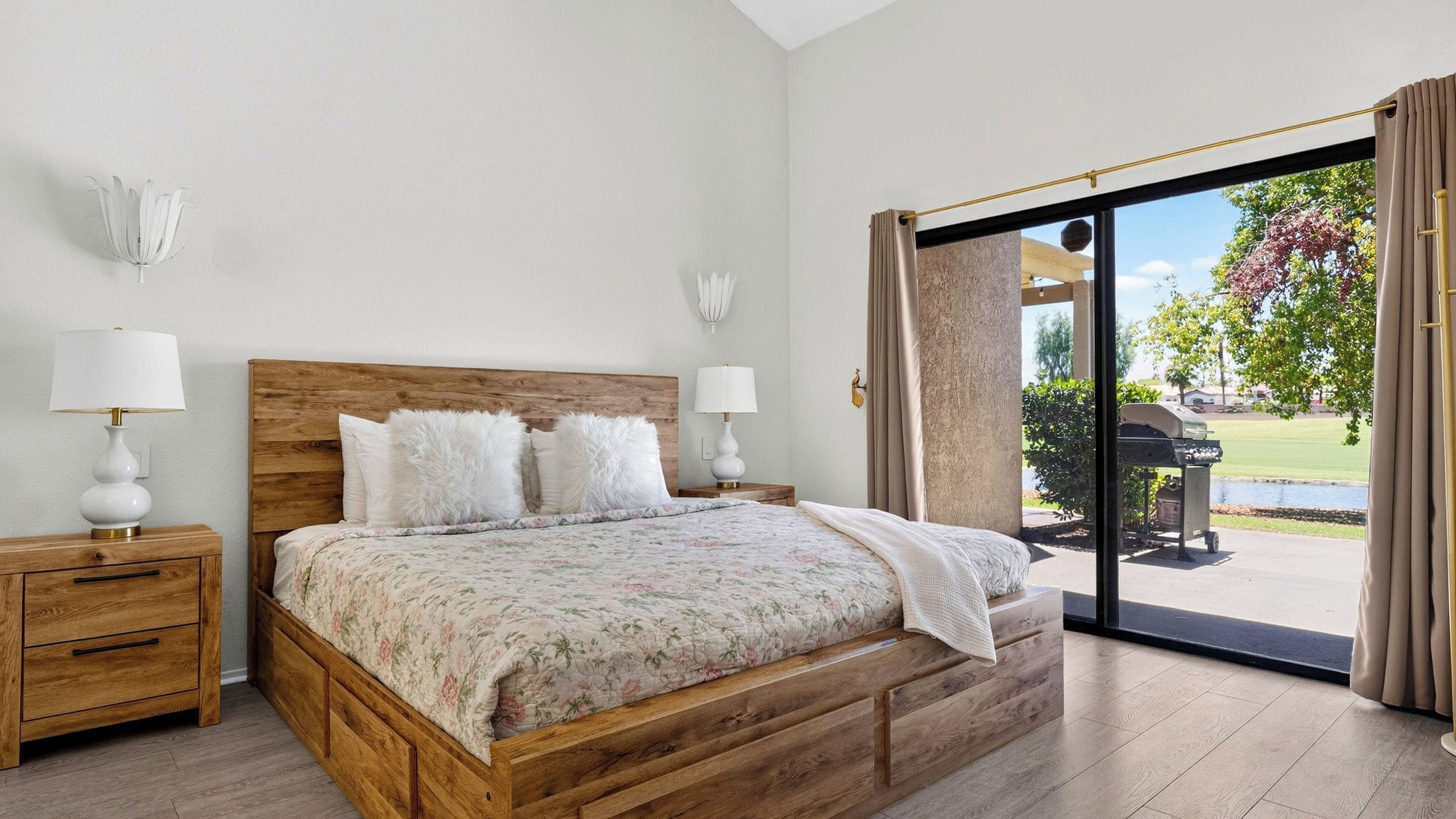 A bedroom with a wooden bed, two bedside tables, a large window with a view of a patio and outdoor grill.