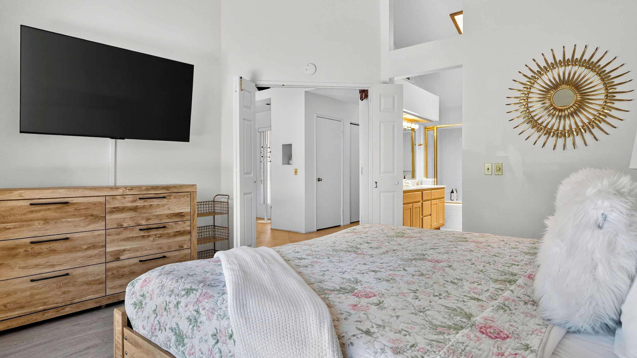 Un dormitorio con cama, televisor, cómoda, espejo y puerta que da a otra habitación.
