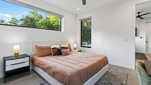 Un dormitorio moderno con una cama grande, dos mesitas de noche con lámparas, vistas a zonas verdes a través de ventanas y un ventilador de techo.