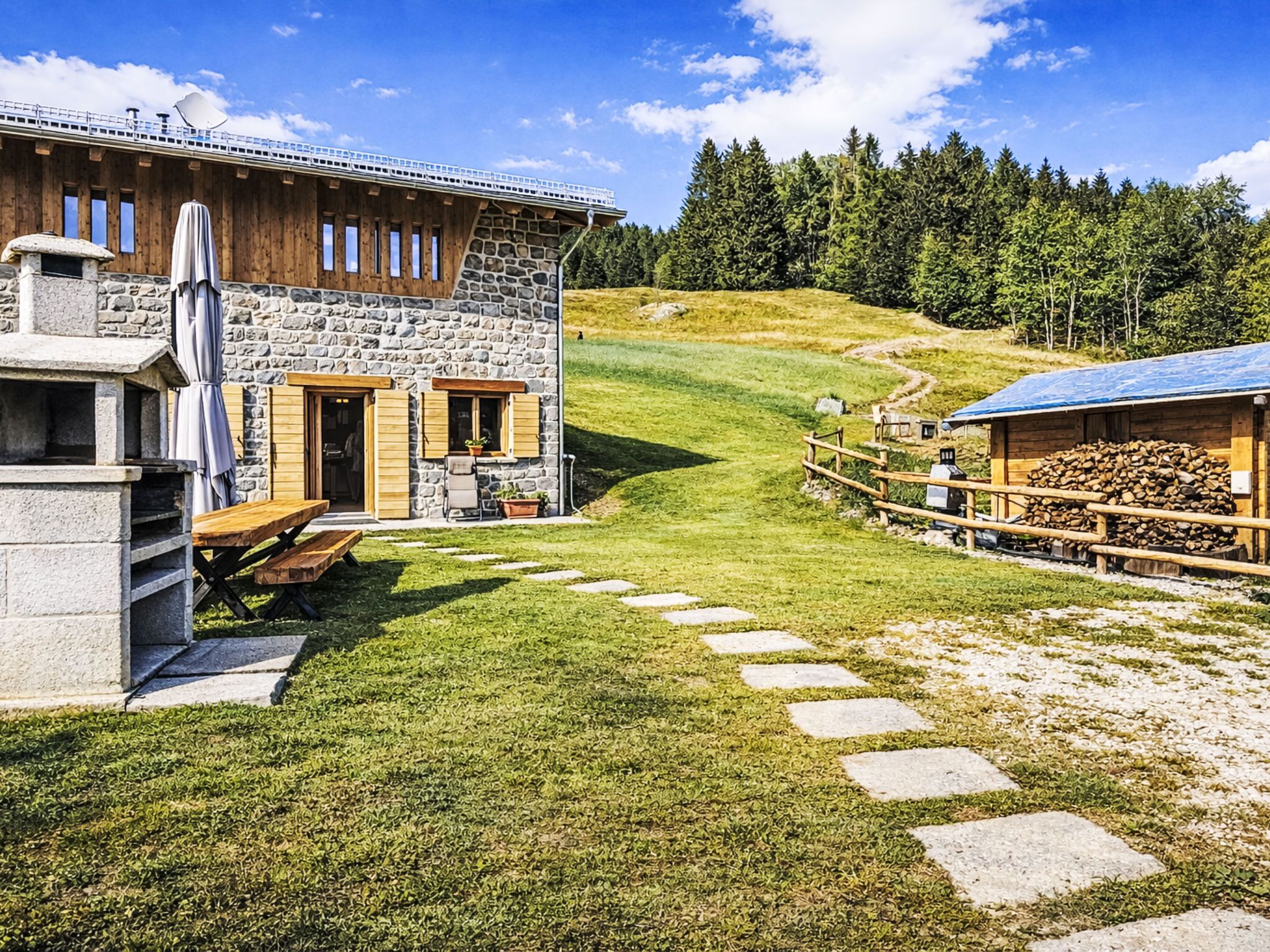 Un edificio in pietra con un patio in legno e una panchina in pietra, immerso in un'area erbosa e circondato da un bosco.