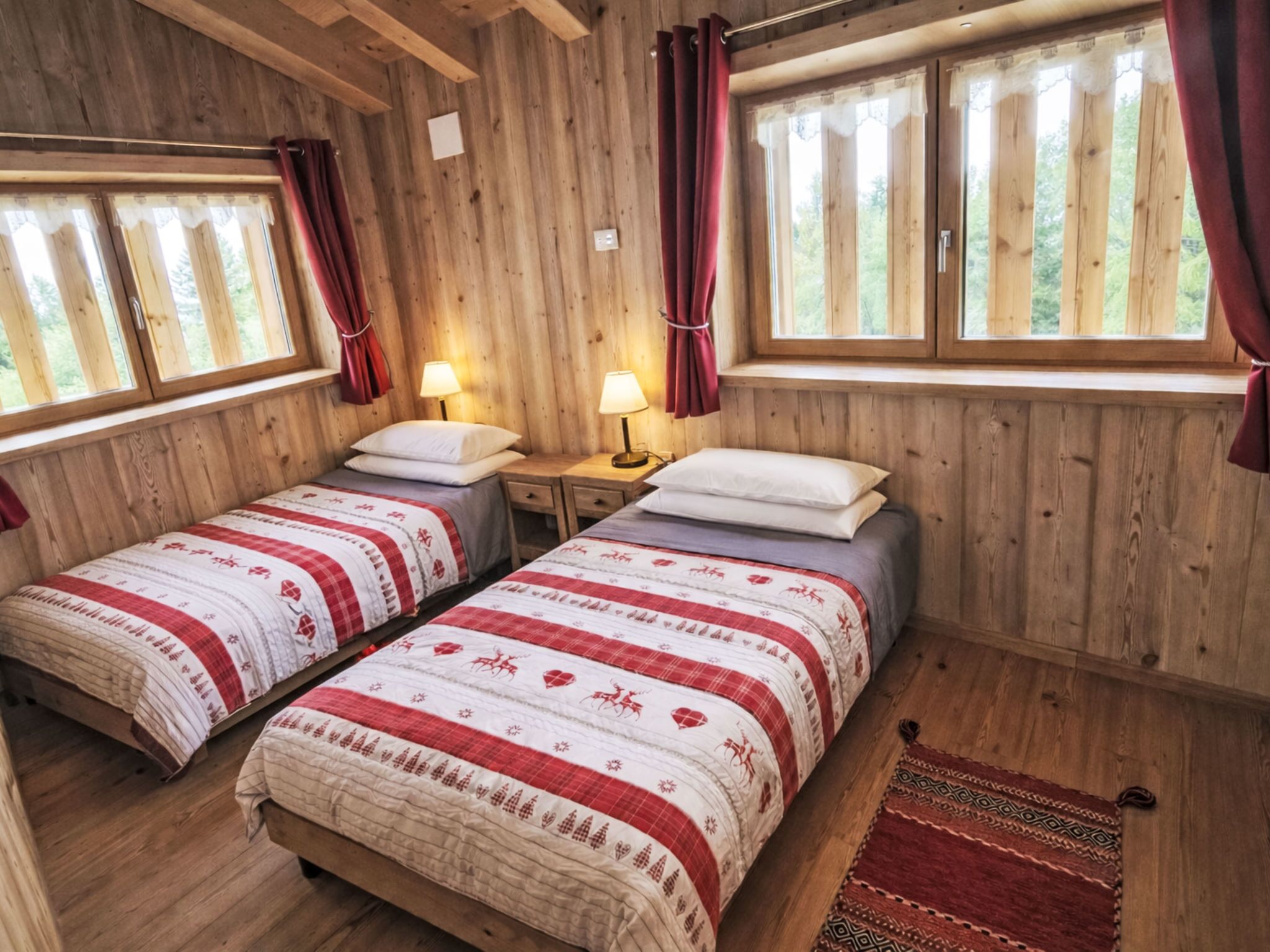 Ein Zimmer in einer Holzhütte mit zwei Betten, jedes mit einer rot-weißen Musterdecke, zwei Nachttischen mit Lampen und großen Fenstern mit Vorhängen.
